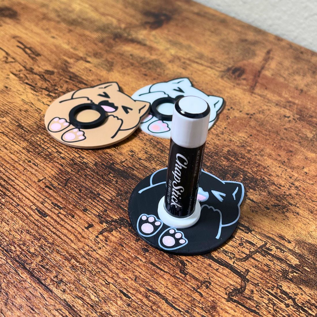 Three chonk cat chapstick stands sitting on wooden tabletop one having a tube of chapstick inside it showing how the product works