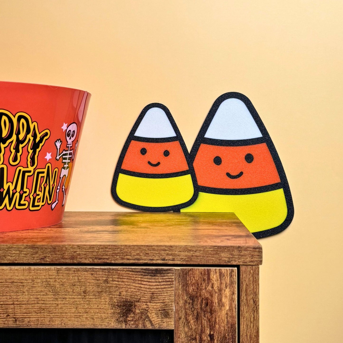 Two happy candy corn door toppers on a rustic cabinet corner next to a Happy Halloween candy bucket