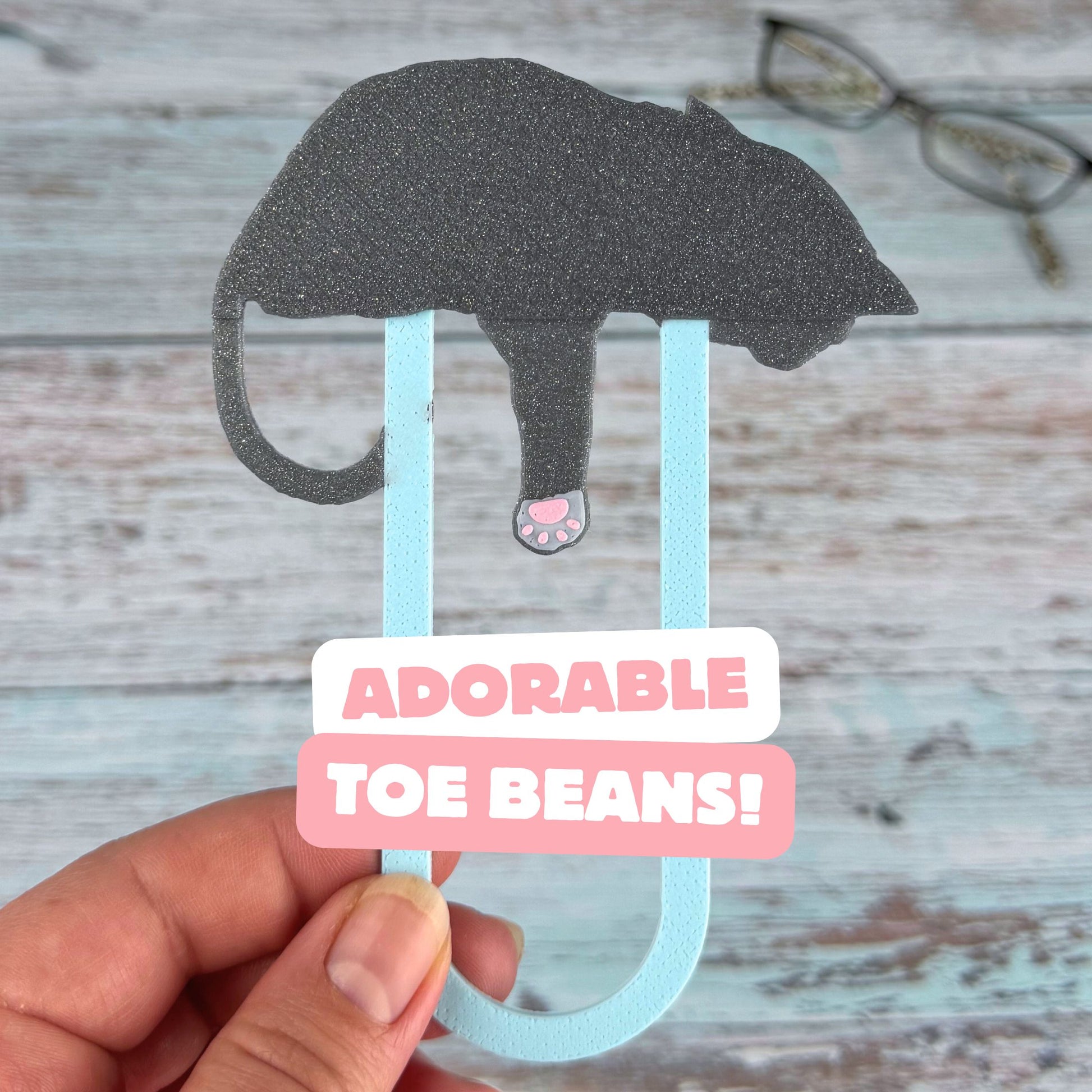 The back of a grey tuxedo cat bookmark showing the adorable pink toe beans on the back of its paw