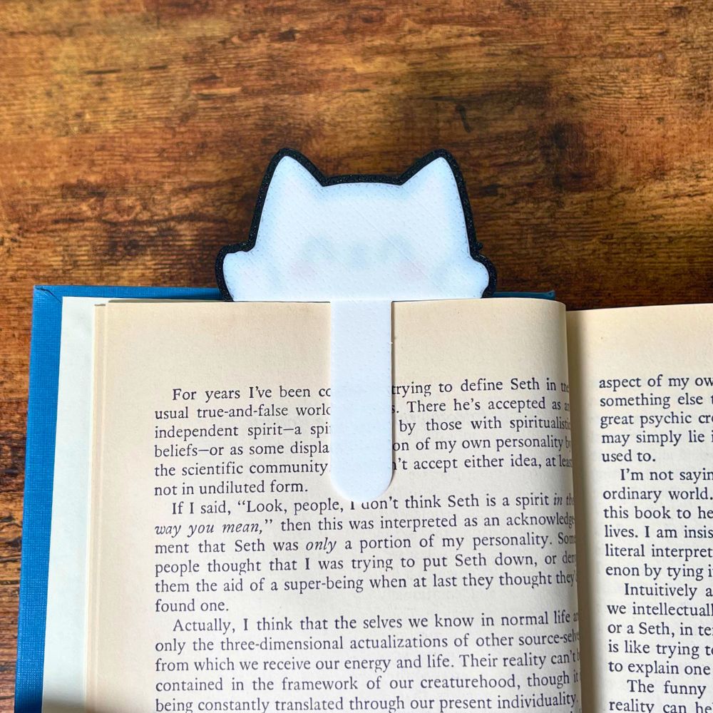 The backside view of the kawaii style cat bookmark clipped onto the page of a blue book from an angle
