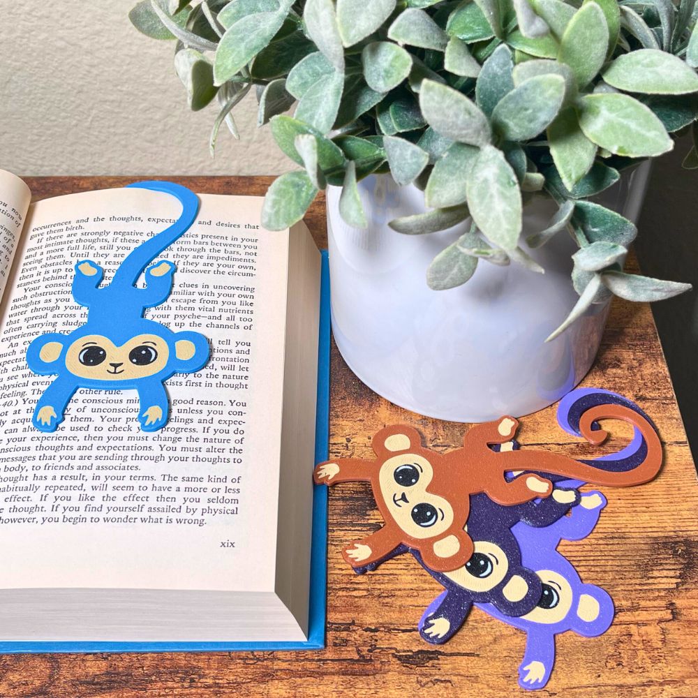 Four colors of fun monkey bookmarks hanging from a book and on a shelf