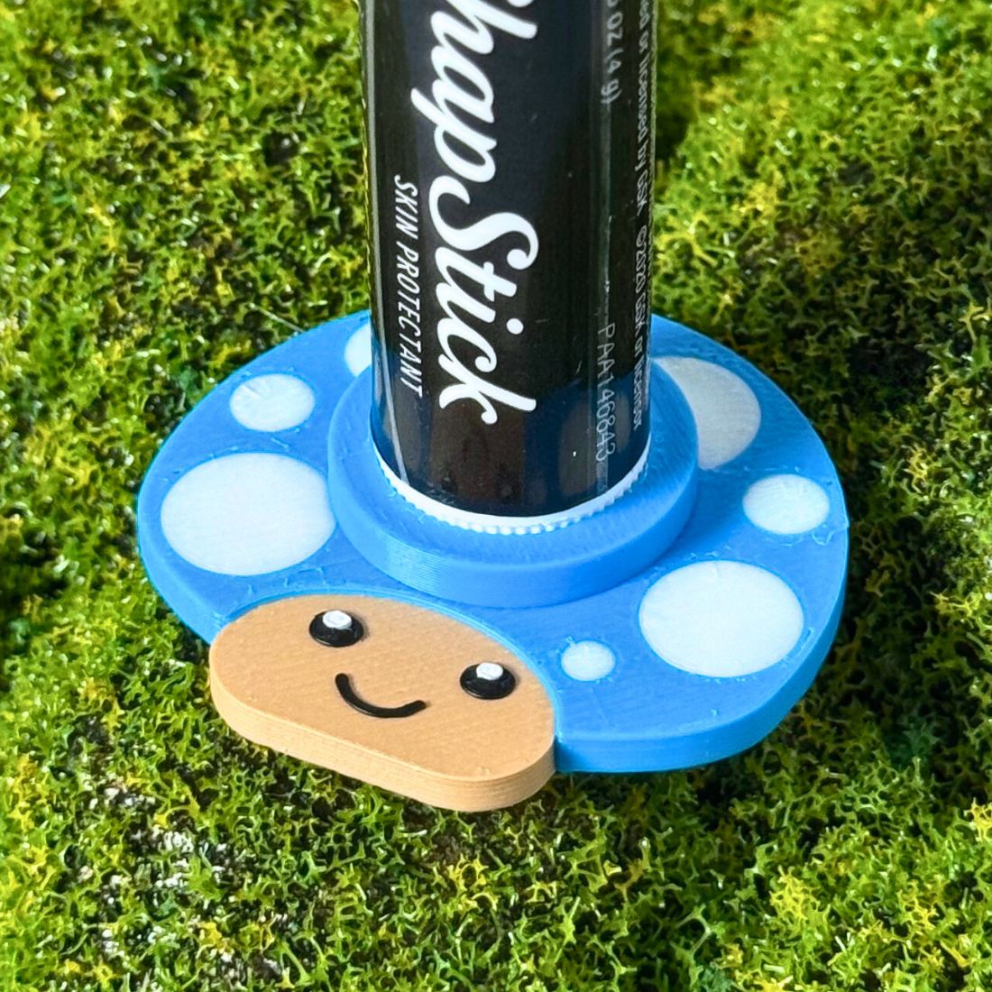 Happy little mushroom lip balm holder in cyan blue on moss holding a chapstick up