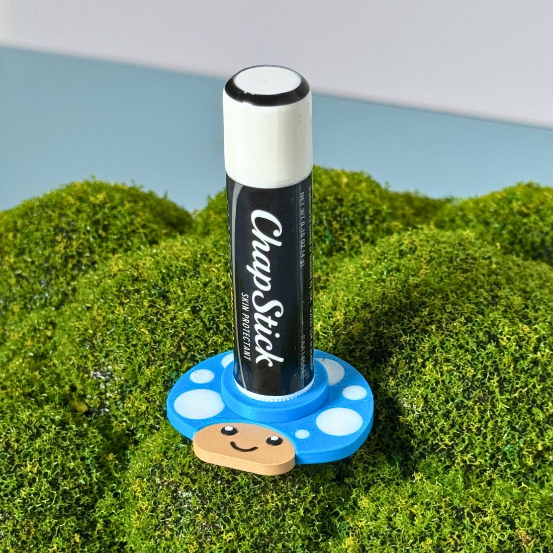 Happy little mushroom lip balm holder in cyan blue on moss holding a chapstick up