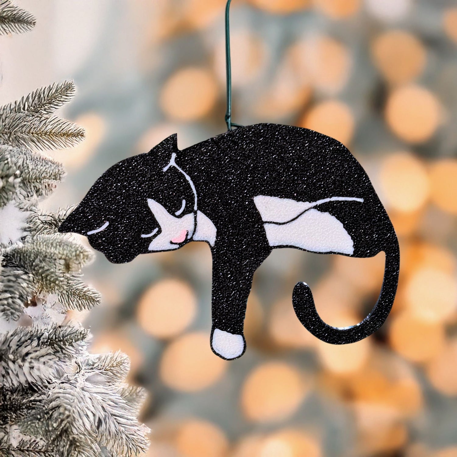 Glittery tuxedo cat ornament hanging beside a frosted tree with golden bokeh lights, a small stocking stuffer