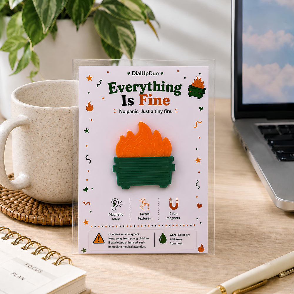 Everything Is Fine dumpster fire magnetic fidget in DialUpDuo packaging on a desk beside a laptop