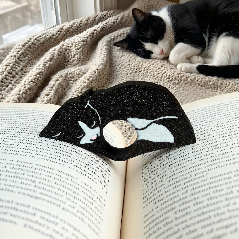 Tuxedo cat-shaped book page holder on an open book with a cat lying on a blanket in the background
