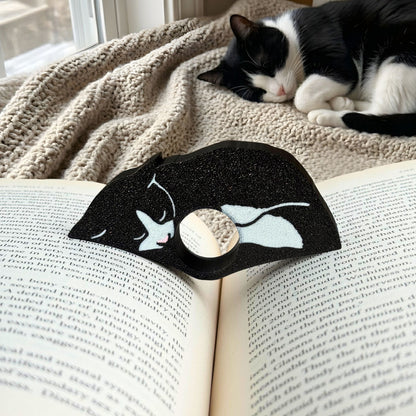 Tuxedo cat-shaped book page holder on an open book with a cat lying on a blanket in the background