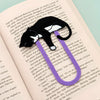 A purple paperclip-shaped bookmark shaped like a black and white tuxedo cat, resting on open book pages.