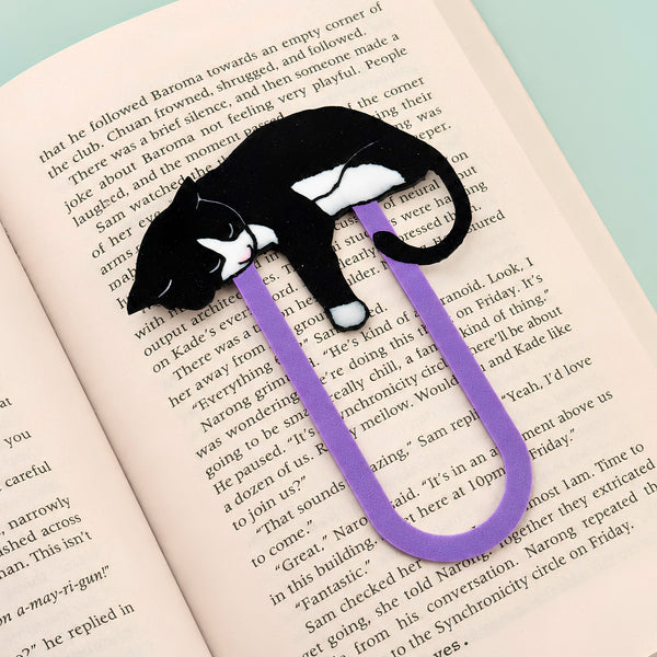 A purple paperclip-shaped bookmark shaped like a black and white tuxedo cat, resting on open book pages.