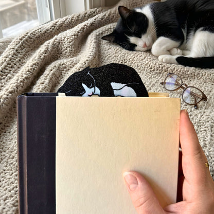 Person holding a closed book with a cat-shaped bookmark, cat lying on a blanket in the background