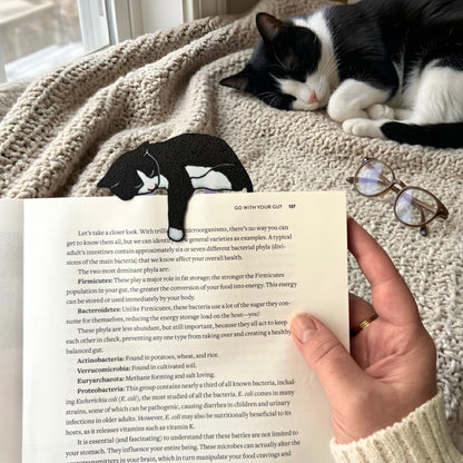 Person holding a book with a tuxedo cat bookmark, cat lying on a blanket in the background