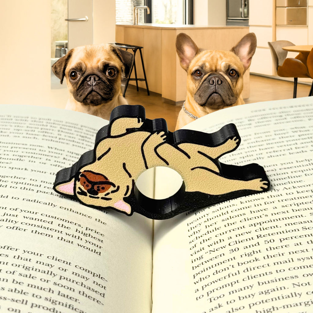 Adorable tan bulldog page holder resting across an open book with two curious pug and French bulldog dogs in the background, showcasing a cute and functional reading accessory for book lovers.