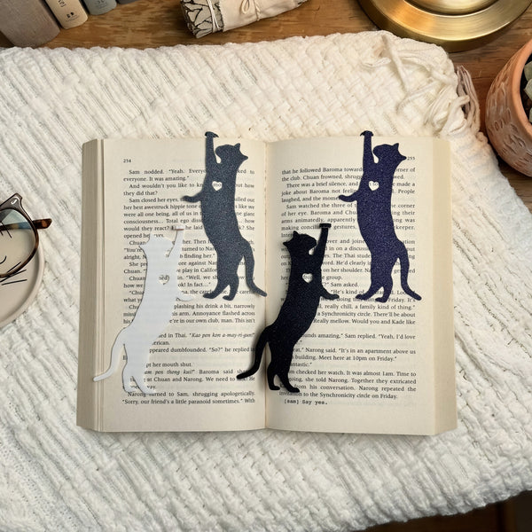 Cat hanging by paw bookmarks in white, navy and black stretching down the pages of an open book