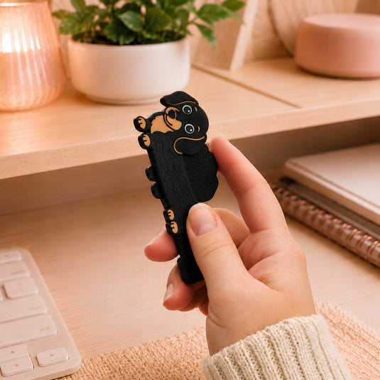 Black and tan dachshund magnetic fidget held in one hand at a cozy desk, showing the textured dog-shaped sensory accessory in use beside a keyboard, notebook, plant, and warm lamp.