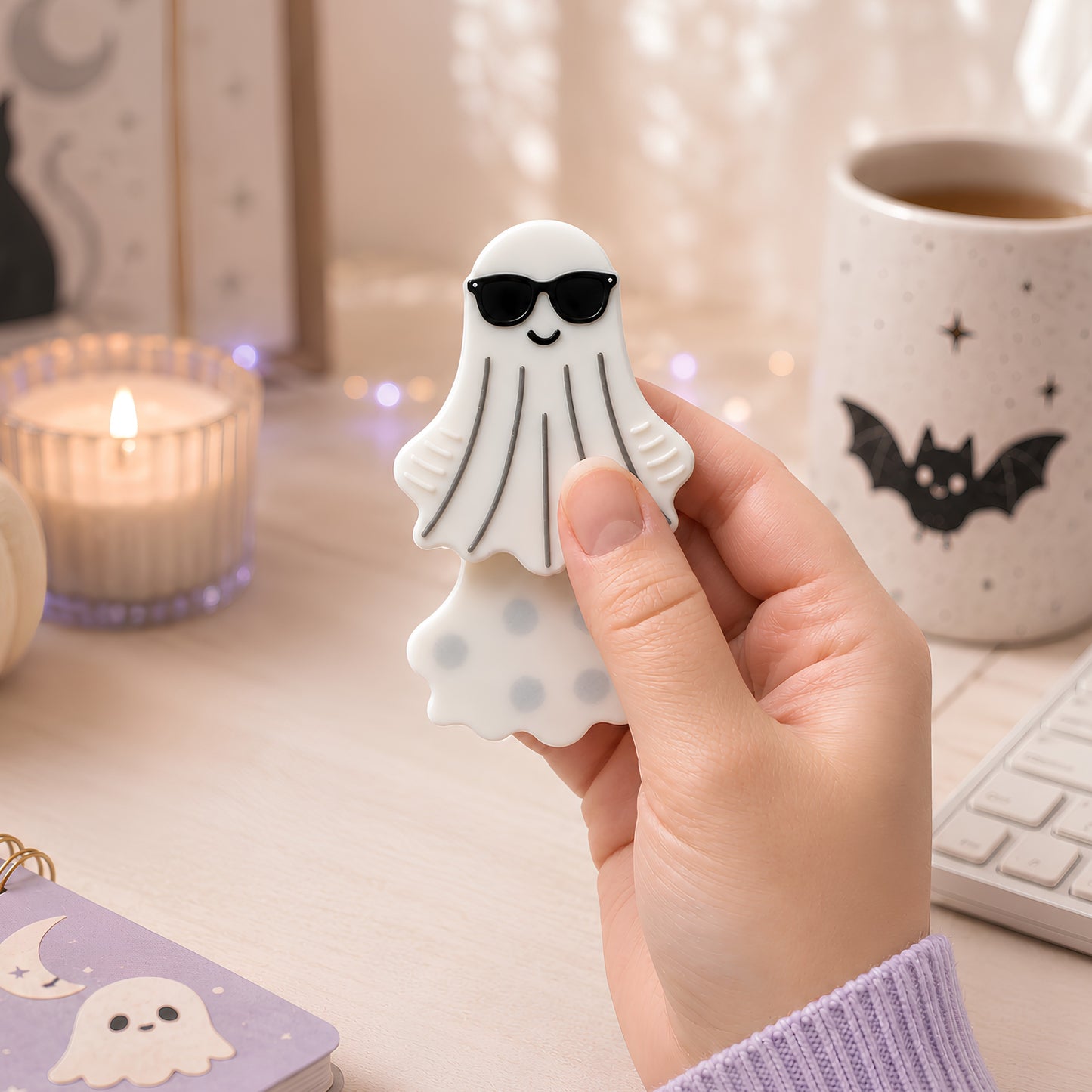 A hand holding a white ghost-shaped magnetic fidget with sweet black sunglasses, set against a cozy desk background with a lit candle, a mug, and a keyboard.