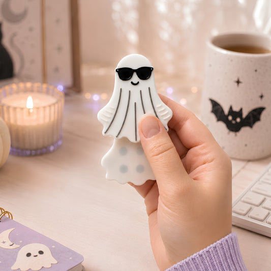 A hand holding a white ghost-shaped magnetic fidget with sweet black sunglasses, set against a cozy desk background with a lit candle, a mug, and a keyboard.
