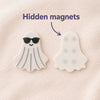 Two ghost-shaped items on a soft off-white blanket textured background, with text reading 'Hidden magnets' and an arrow pointing to the back of the ghost fidget on the right showing the magnet pattern. The front side of the fidget on the left shows black sunglasses and has gray tactile lines.