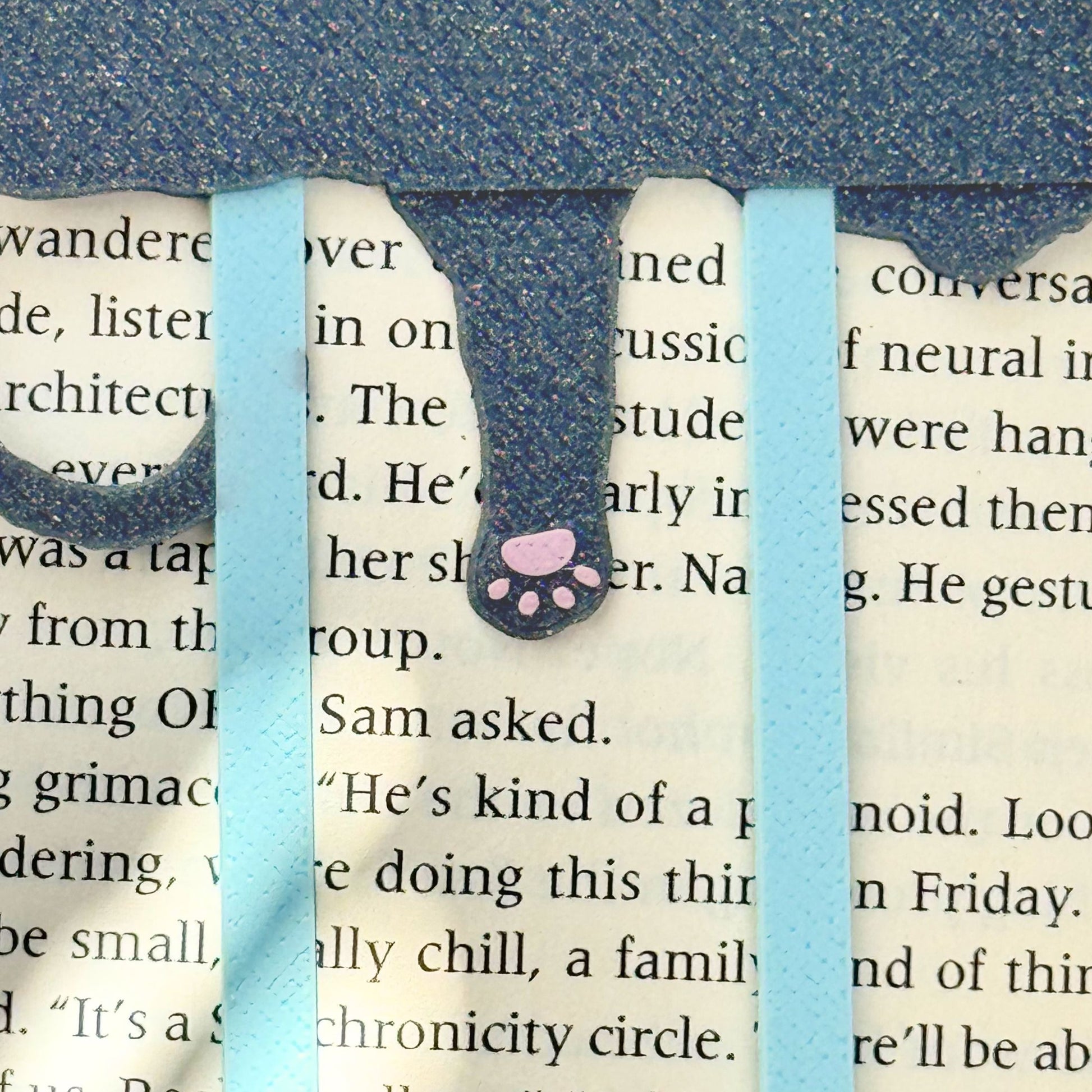 Back view of the bookmark showing a cat paw with pink toe beans with a light blue clip on an open book