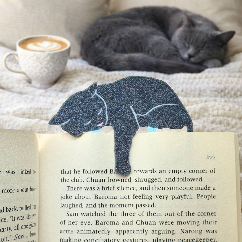 Gray cat-shaped bookmark on an open book with a cat and coffee cup in the background