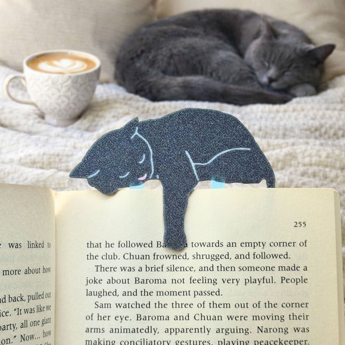 Gray cat-shaped bookmark on an open book with a cat and coffee cup in the background