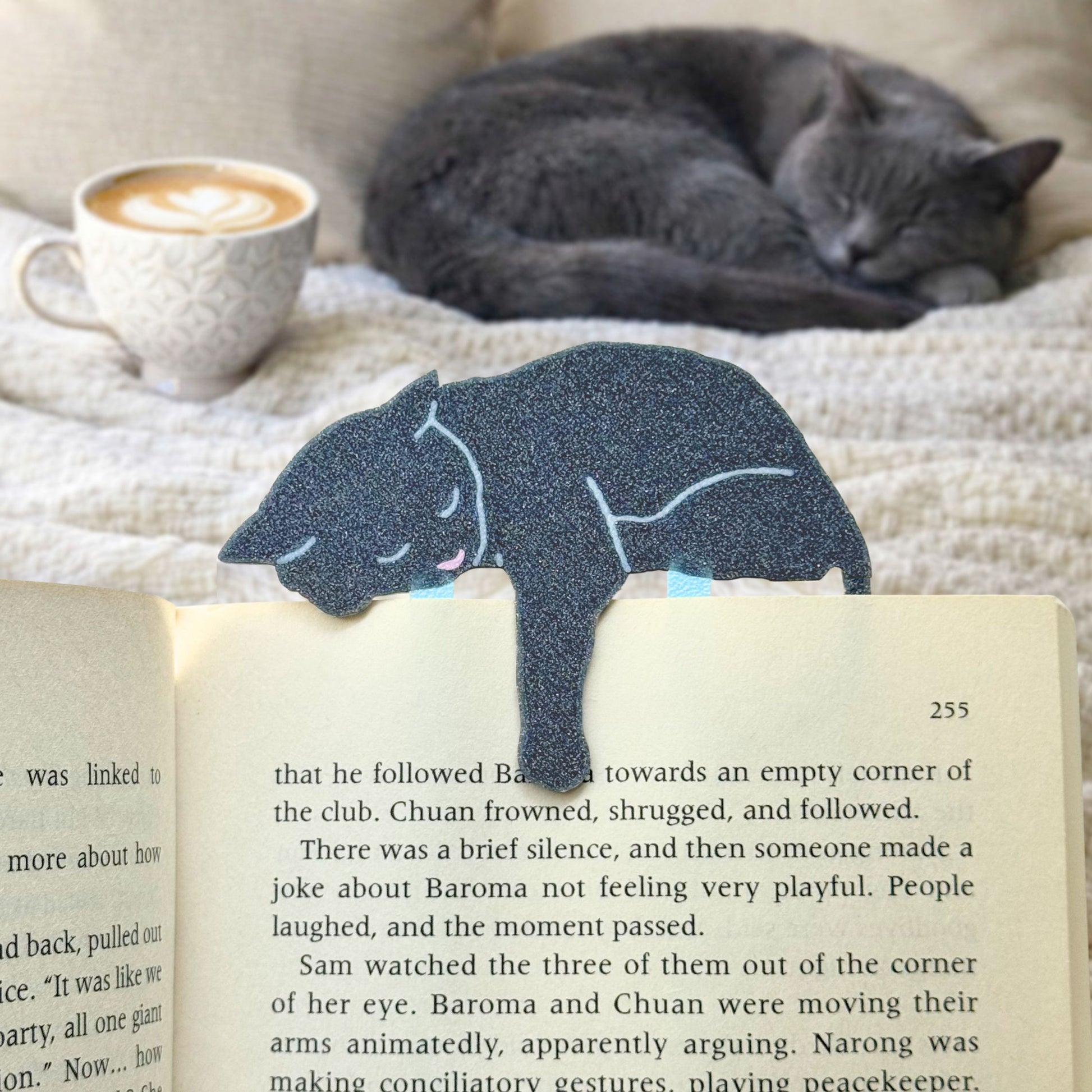 Gray cat-shaped bookmark on an open book with a cat and coffee cup in the background