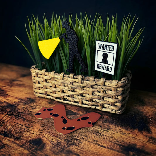 Wicker basket plant with a black silhouette investigator plant stake, a 'Wanted' poster plant stake, and a brown path of footprints on a wooden surface.