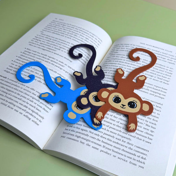 Three fun monkey bookmarks in blue, dark purple and brown hanging from curly tails on an open book