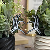 Skeletal hand heart gesture plant stakes sticking out between two potted plants with a blurred indoor background.
