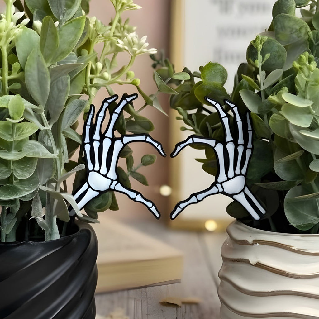 Skeletal hand heart gesture plant stakes sticking out between two potted plants with a blurred indoor background.