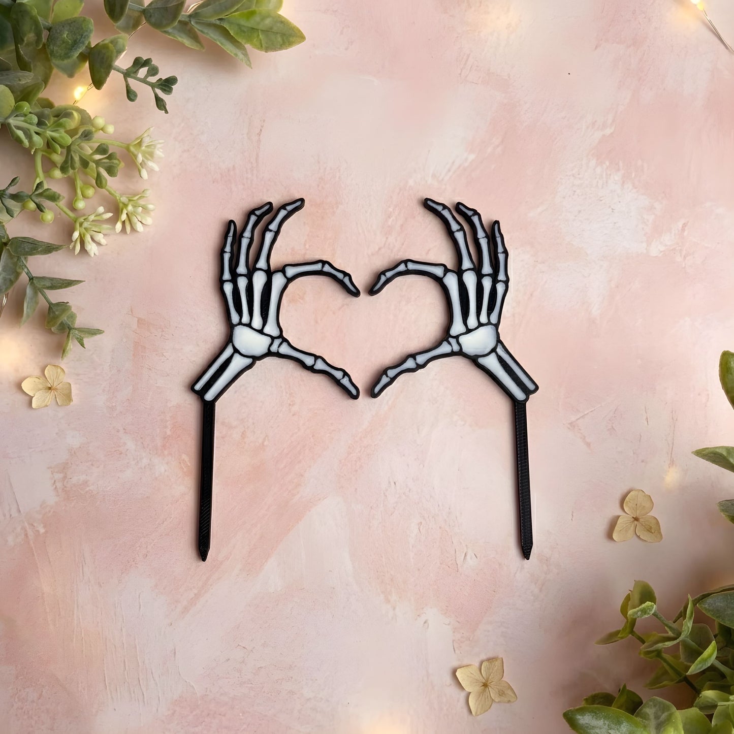 Skeleton hands forming a heart shape on a pink background with greenery