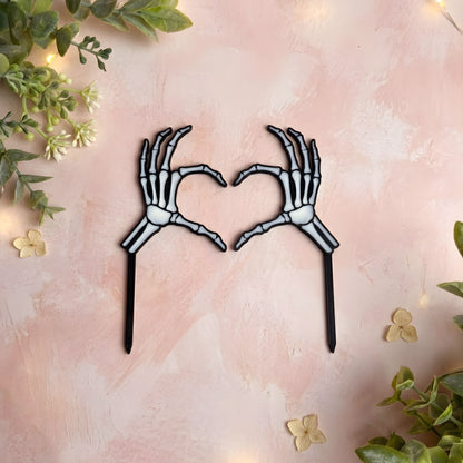Skeleton hands forming a heart shape on a pink background with greenery