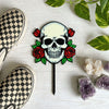 Skull and roses plant stake on a white surface with checkered shoes, plant leaves, and crystals surrounding the stake.