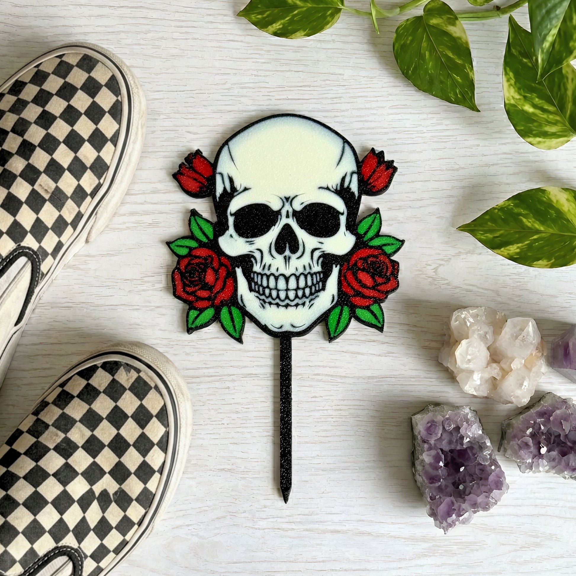 Skull and roses plant stake on a white surface with checkered shoes, plant leaves, and crystals surrounding the stake.