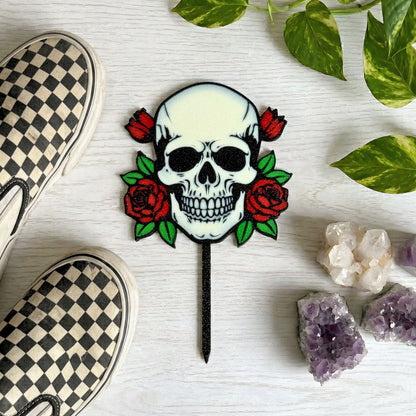 Skull and roses plant stake on a white surface with checkered shoes, plant leaves, and crystals surrounding the stake.