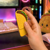 Yellow taco magnetic fidget held partially open in one hand at a colorful RGB gaming desk, showing the raised shell texture, layered toppings, and full sliding base beside a glowing keyboard, mouse, and taco plush.