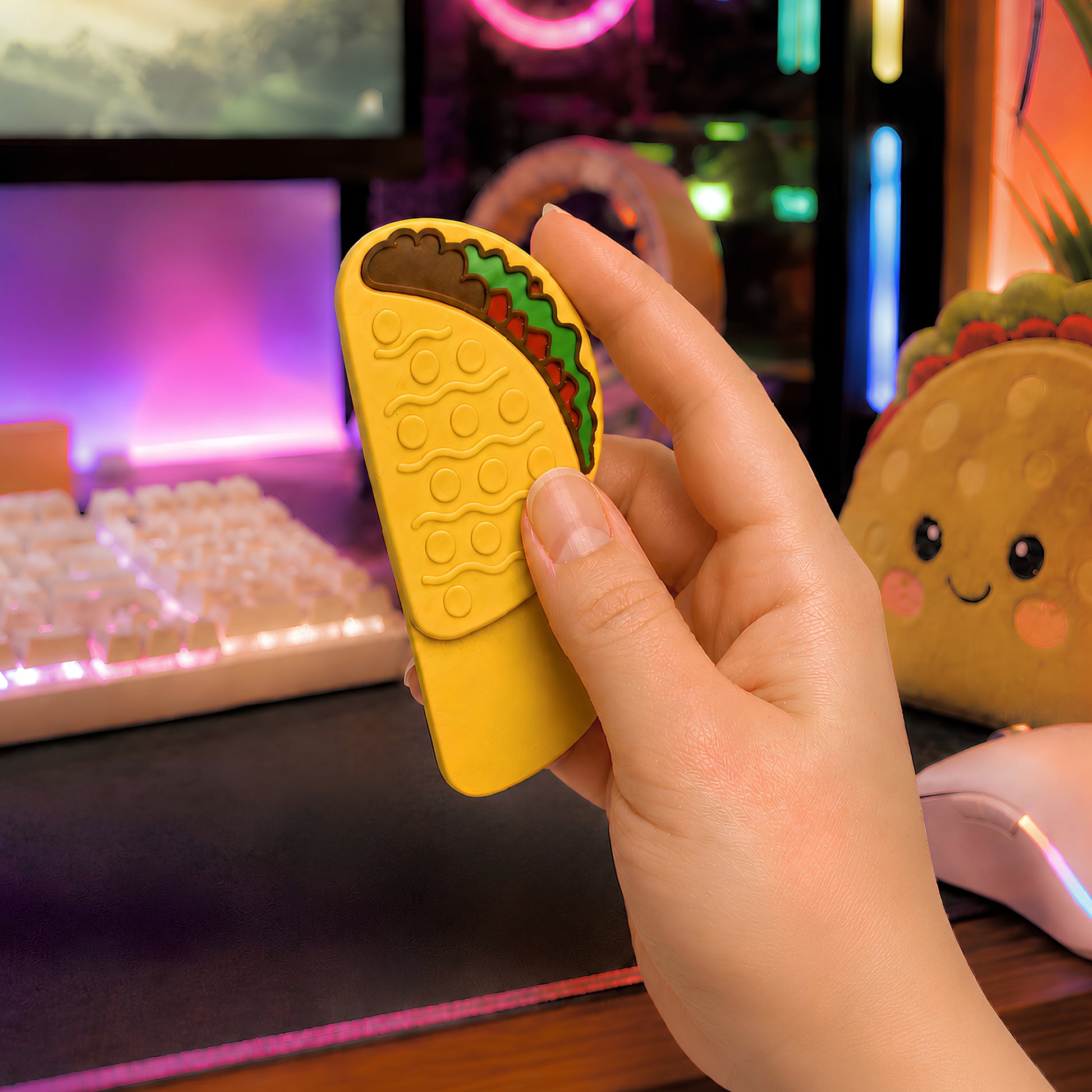Yellow taco magnetic fidget held partially open in one hand at a colorful RGB gaming desk, showing the raised shell texture, layered toppings, and full sliding base beside a glowing keyboard, mouse, and taco plush.