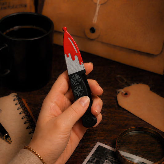 Red, silver, and black knife-shaped magnetic fidget held in one hand on a moody true crime desk with a fingerprints on the handle, black mug, notebook, evidence tag, and magnifying glass.