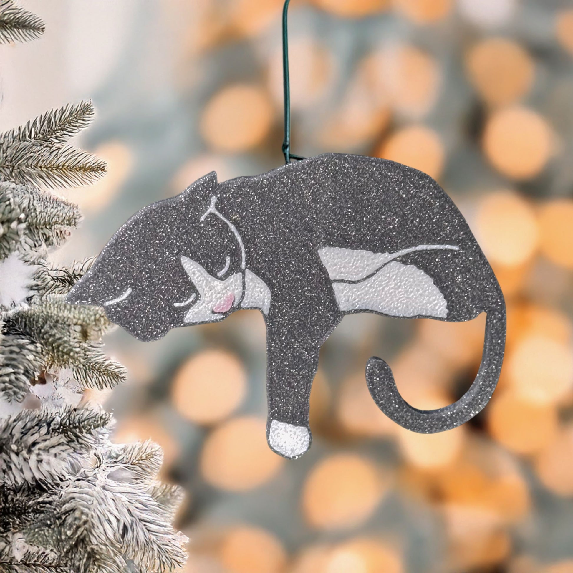 Gray and white tuxedo cat ornament with pink toe beans hanging on a frosted Christmas tree branch, surrounded by warm holiday lights, perfect gift for cat parents and festive home decor