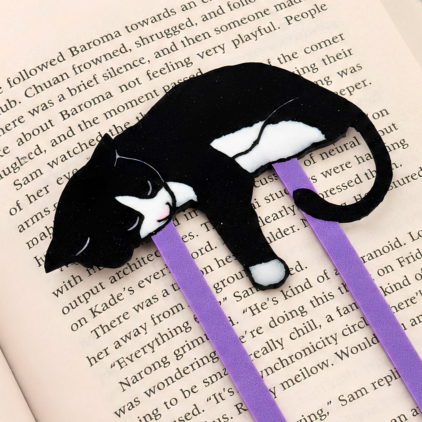 A black and white tuxedo cat-shaped bookmark with purple paperclip, resting on an open book page.