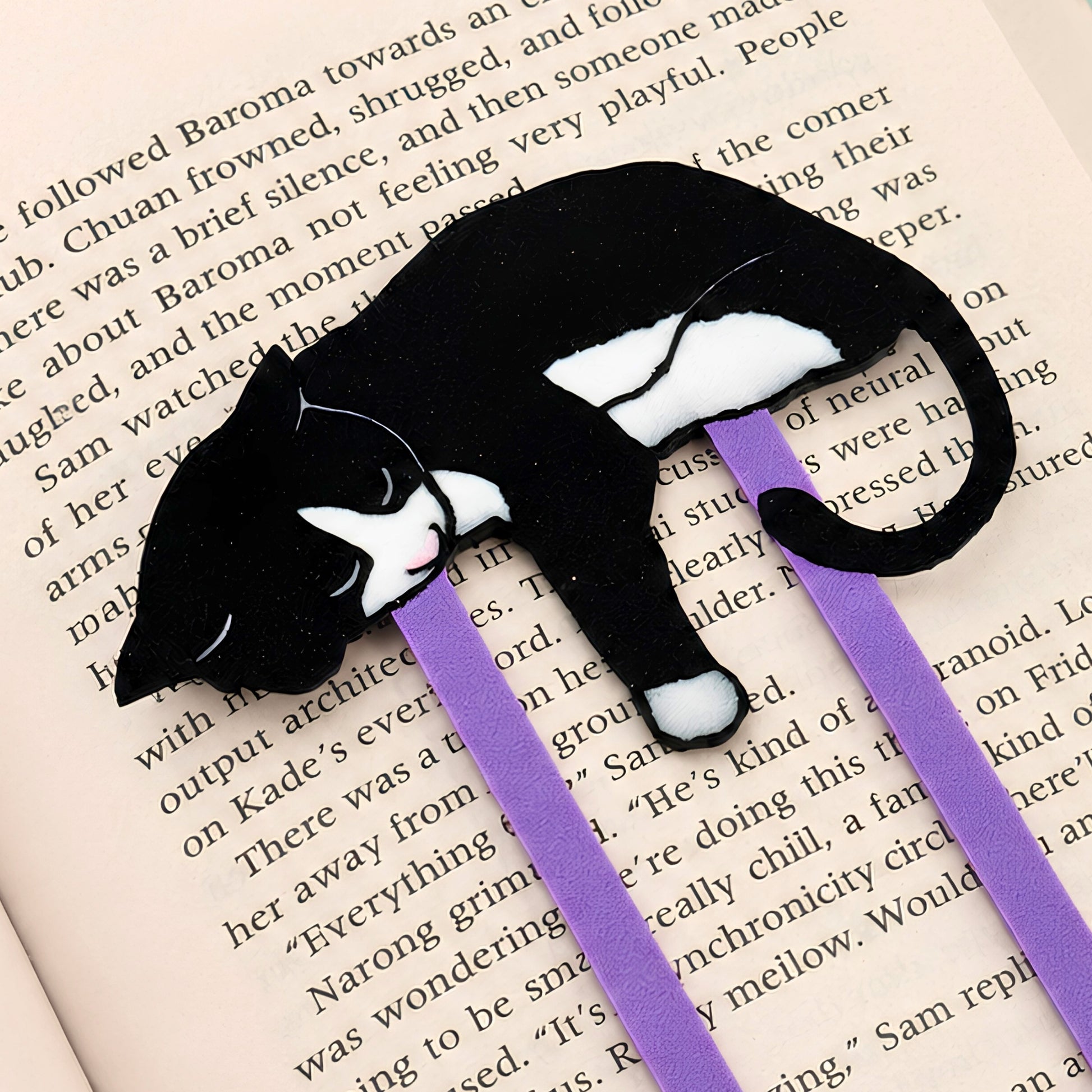 A black and white tuxedo cat-shaped bookmark with purple paperclip, resting on an open book page.
