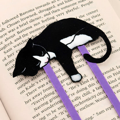 A black and white tuxedo cat-shaped bookmark with purple paperclip, resting on an open book page.