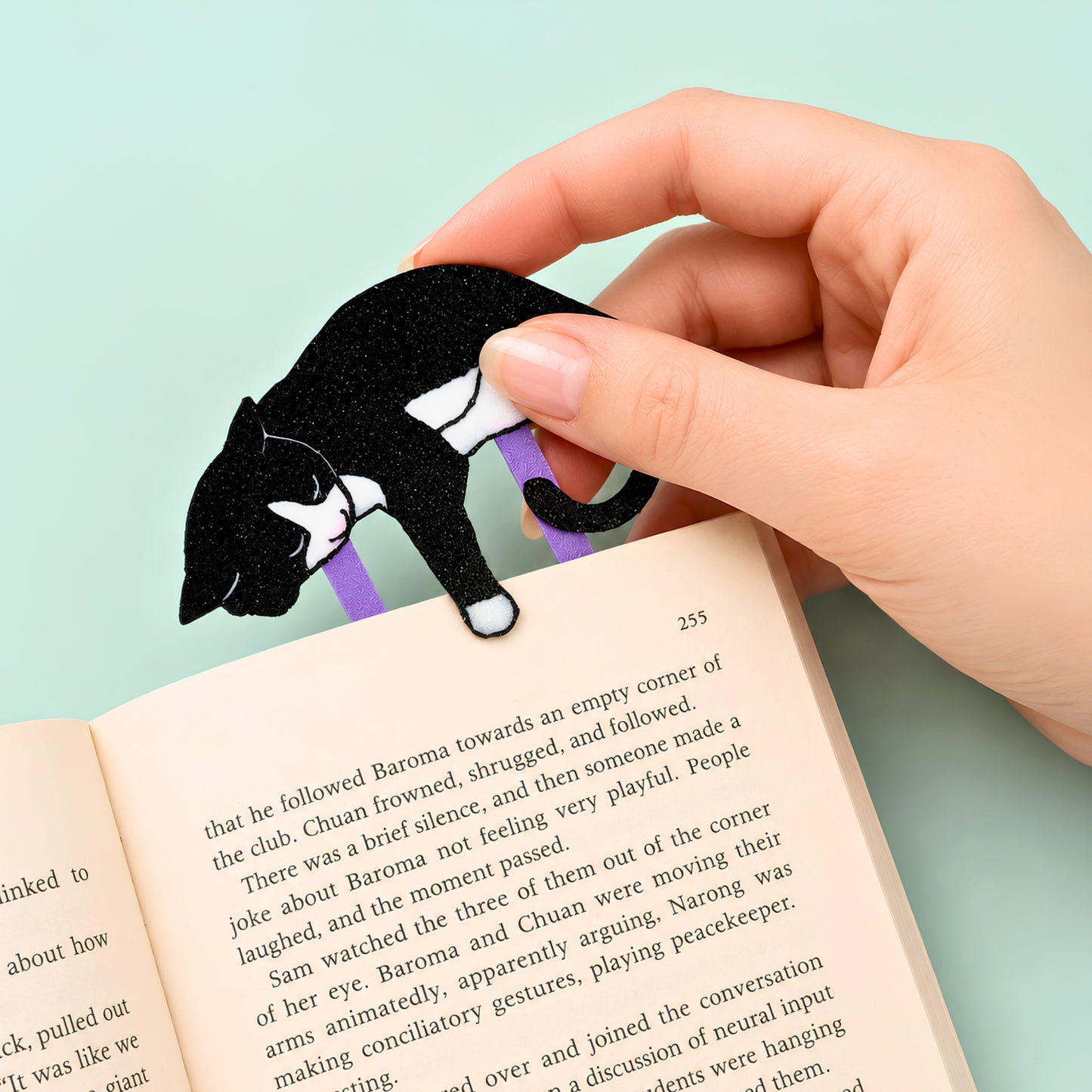 A hand holds a black and white tuxedo cat-shaped bookmark with purple clip in an open book against a light teal green background.