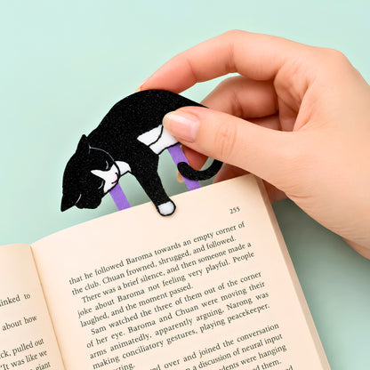 A hand holds a black and white tuxedo cat-shaped bookmark with purple clip in an open book against a light teal green background.
