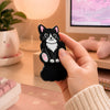 Black and white tuxedo cat magnetic fidget held in one hand at a pastel pink office desk, showing the cat-shaped sensory toy in use beside a keyboard, monitor, plant, and warm lamp.