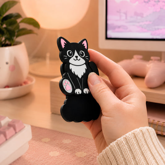 Black and white tuxedo cat magnetic fidget held in one hand at a pastel pink office desk, showing the cat-shaped sensory toy in use beside a keyboard, monitor, plant, and warm lamp.
