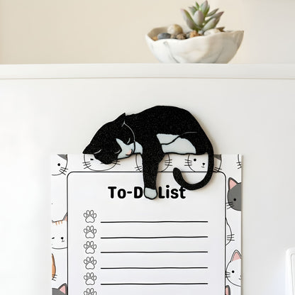 Black tuxedo cat fridge magnet holding a cat-themed to-do list on a refrigerator