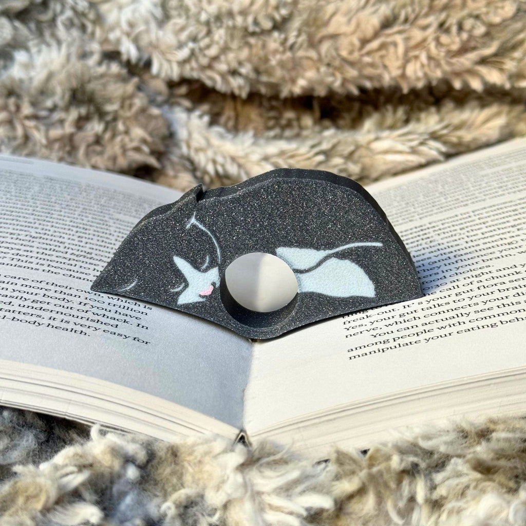 Gray and white cat-shaped thumb book holder ring on an open book with a blanket in the background