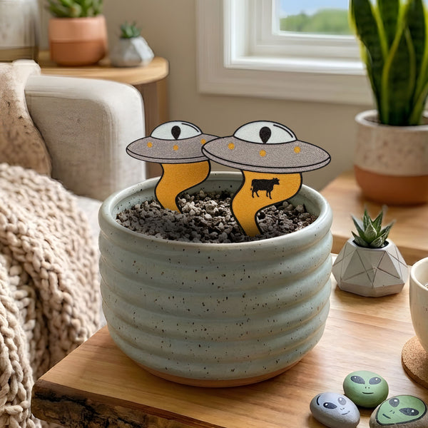 Two alien UFO plant stakes with yellow tractor beams, one abducting a cow, in a speckled indoor planter
