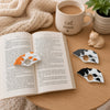 Two white spotted cats in black and gray are displayed on a wooden table next to an open book with the orange and white spotted cat, a ceramic mug with 'cat mom' printed on it, and a small cat figurine.