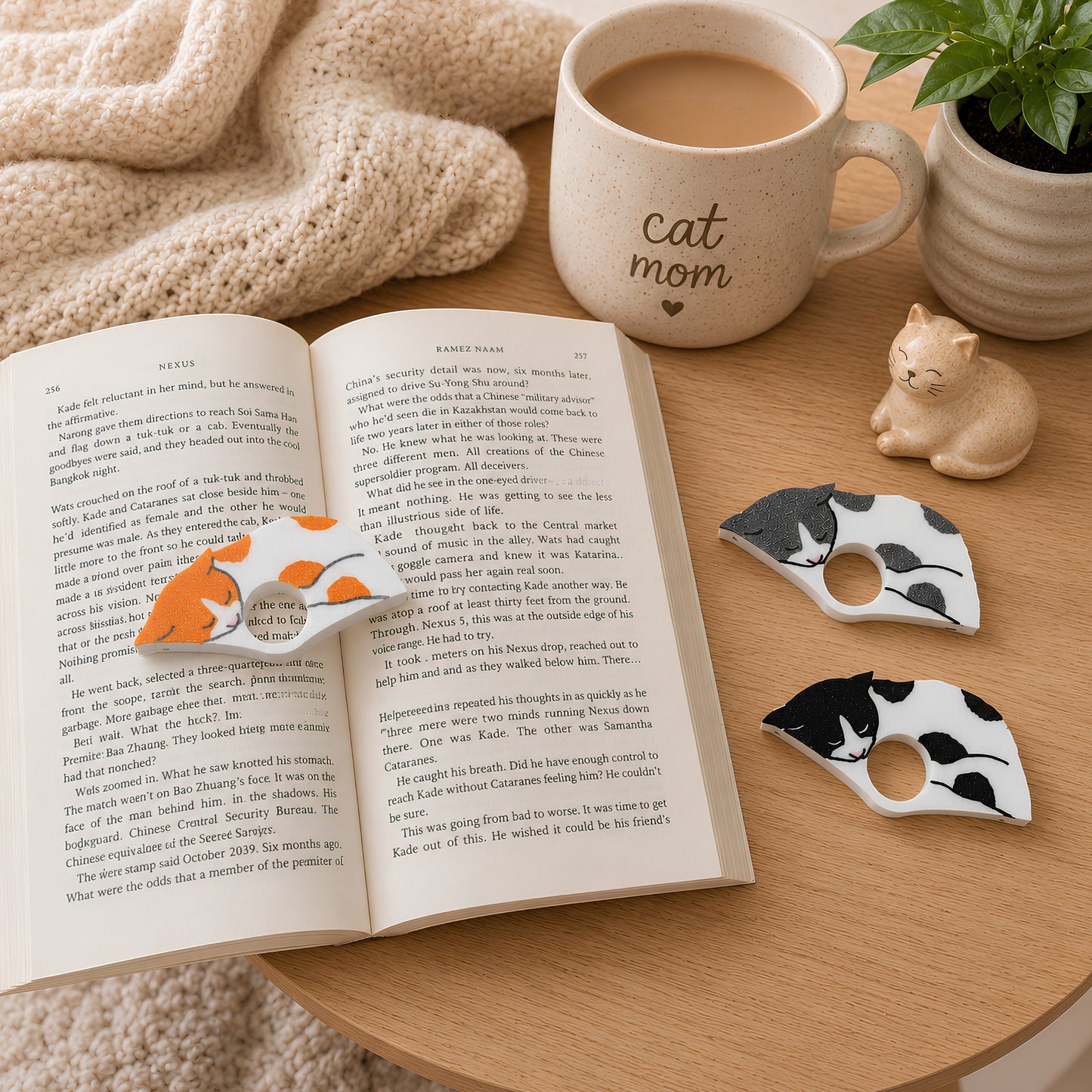 Two white spotted cats in black and gray are displayed on a wooden table next to an open book with the orange and white spotted cat, a ceramic mug with 'cat mom' printed on it, and a small cat figurine.